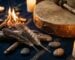 Un tambour chamanique en peau pres d un petit feu, quelques plumes et bois flotte sur une etoffe sombre, tons terre et or