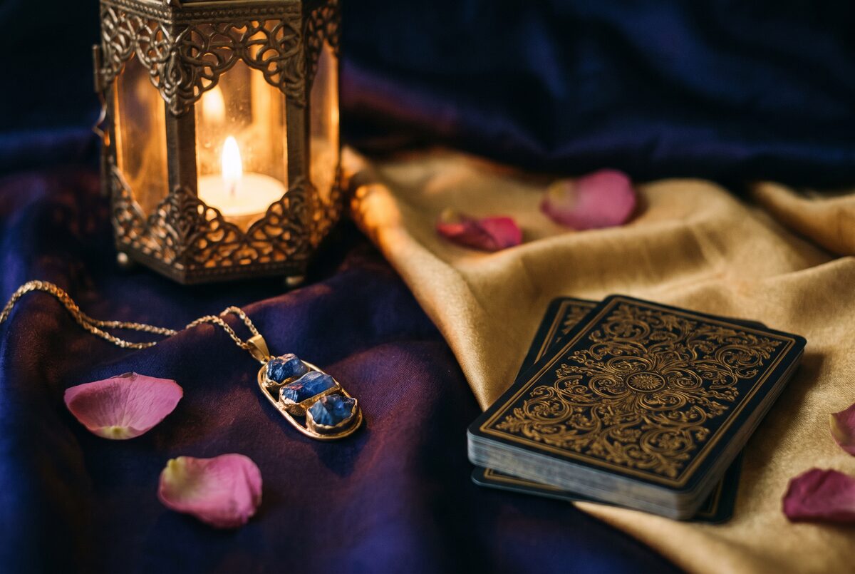 Ambiance romantique à la bougie : jeu de cartes face cachée au dos doré, pétales de rose, pendentif à pierres bleues et lanterne orientale sur des soies bleu nuit et or